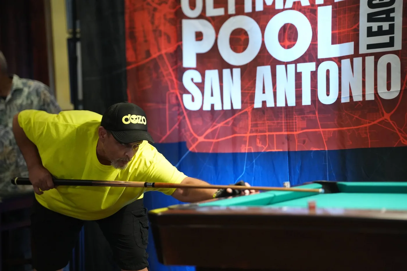 A player in a yellow shirt down on a long shot in front of the Ultimate Pool League San Antonio banner during the 10-Ball qualifier.