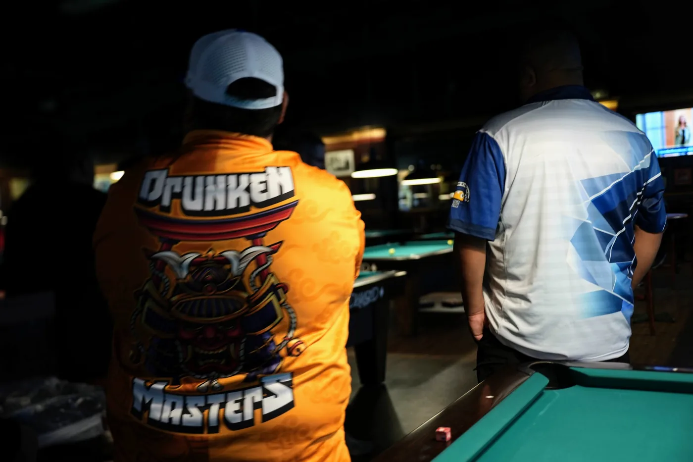 Two players seen from behind at the 10-Ball qualifier: one in an orange Drunken Masters team jersey with the samurai design across the back, the other in a blue and white sublimated jersey.