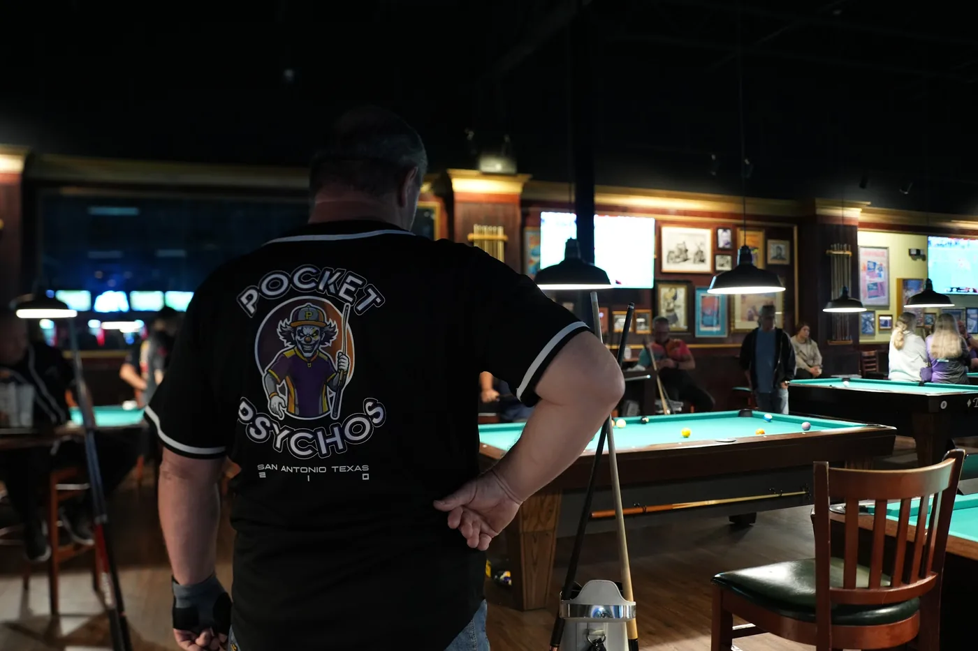A player seen from behind in a black Pocket Psychos baseball-style team jersey, San Antonio Texas under the logo, with play under way across the room.