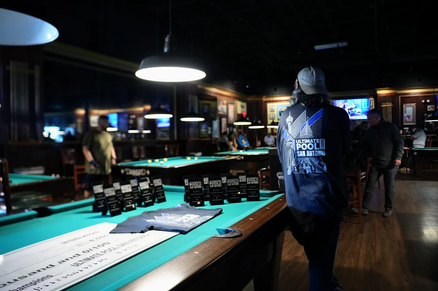 Sunday morning of the 8-Ball qualifier: the champions' and runners-up trophies, the team jerseys and the check laid out on a table under the pendant lights, with play under way across the room behind.